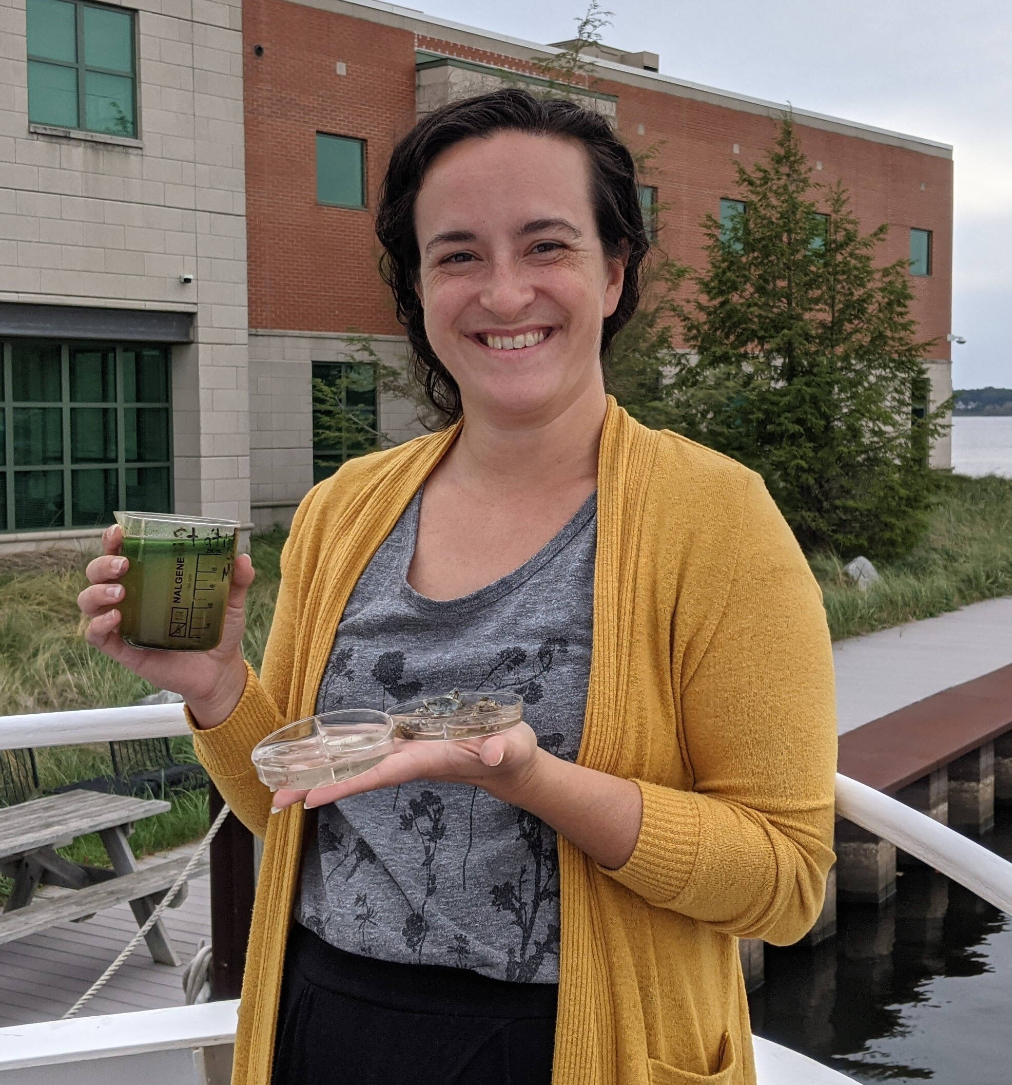 Christina Catanese, outreach program manager, stands on the bow of the W.G. Jackson research vessel. She is smiling and holding a beaker of green liquid (a plankton sample)